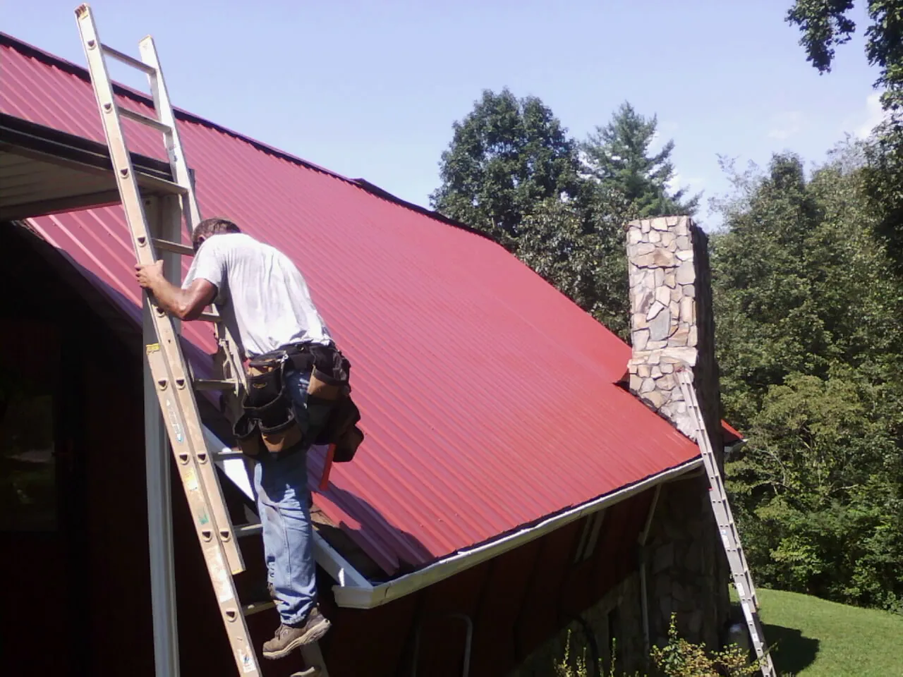 Roofer on a ladder beside a red metal roof and stone chimney — Soffit & Fascia Repair by Roofing Brothers in Granite Canon